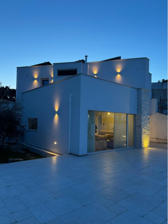 Casa bianca squadrata al crepuscolo con applique accese, vetrata sul soggiorno e cortile pavimentato in pietra chiara